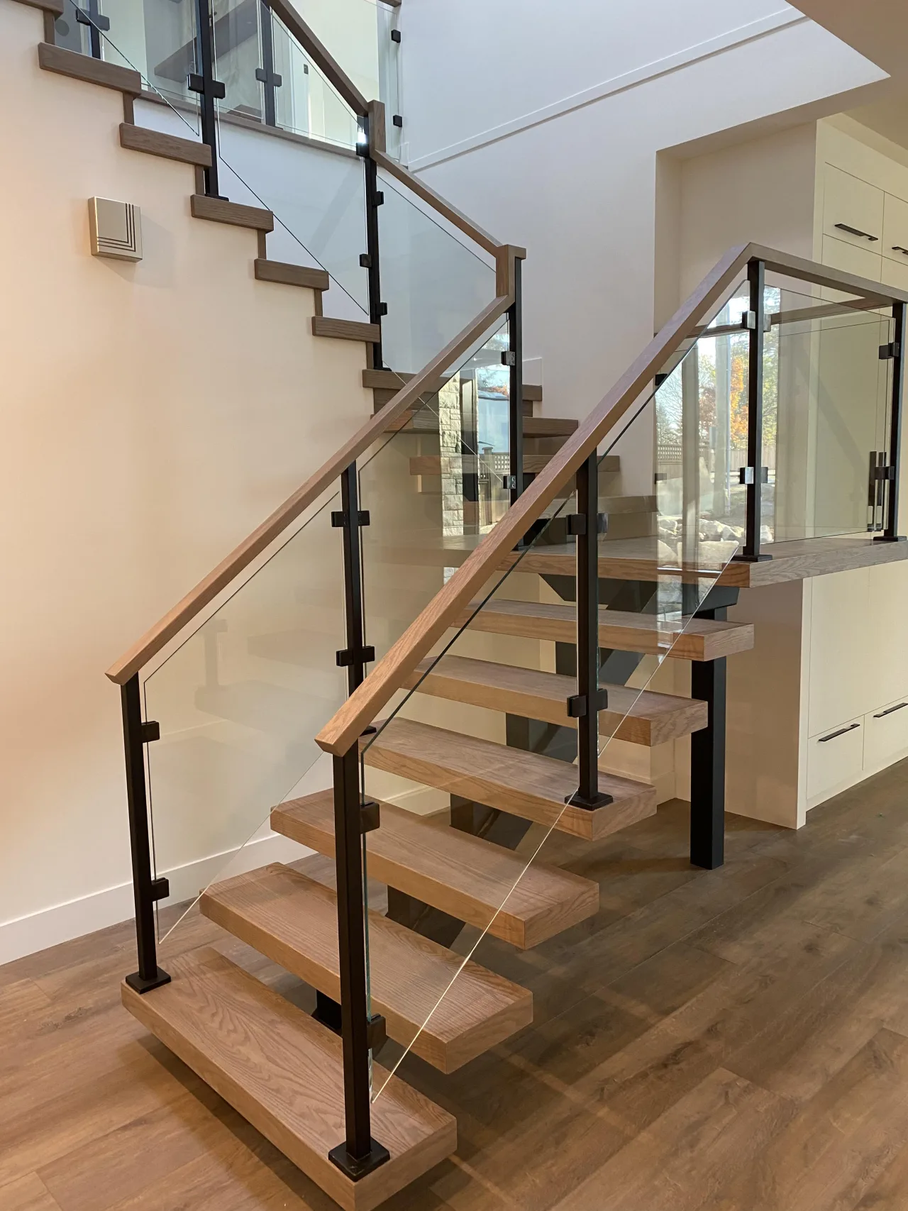 A modern staircase in a home in Coquitlam, BC features wooden steps, a glass railing, and black metal supports.