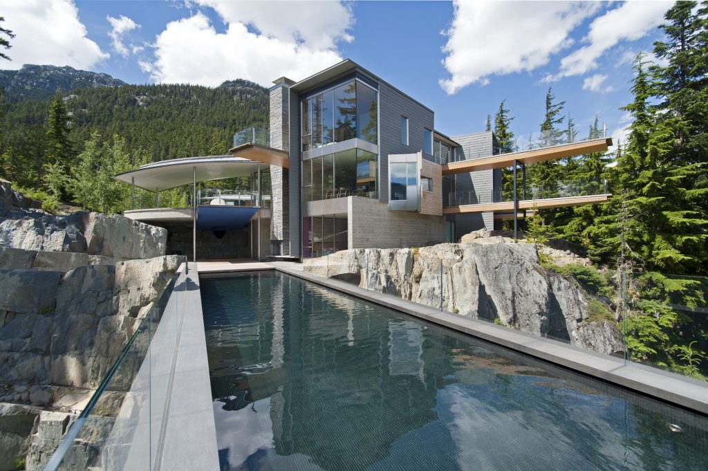Modern architectural home in Coquitlam, BC, features large windows and connecting sky bridges overlooking a reflective pool, all set against a mountainous, forested backdrop.