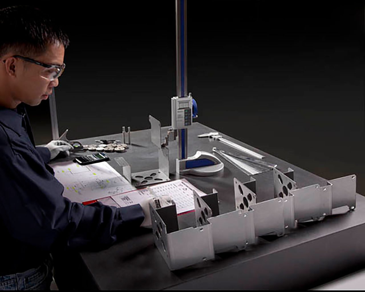 A worker in Coquitlam BC is reviewing technical drawings at a workstation with a digital height gauge, calipers, and manufactured parts.