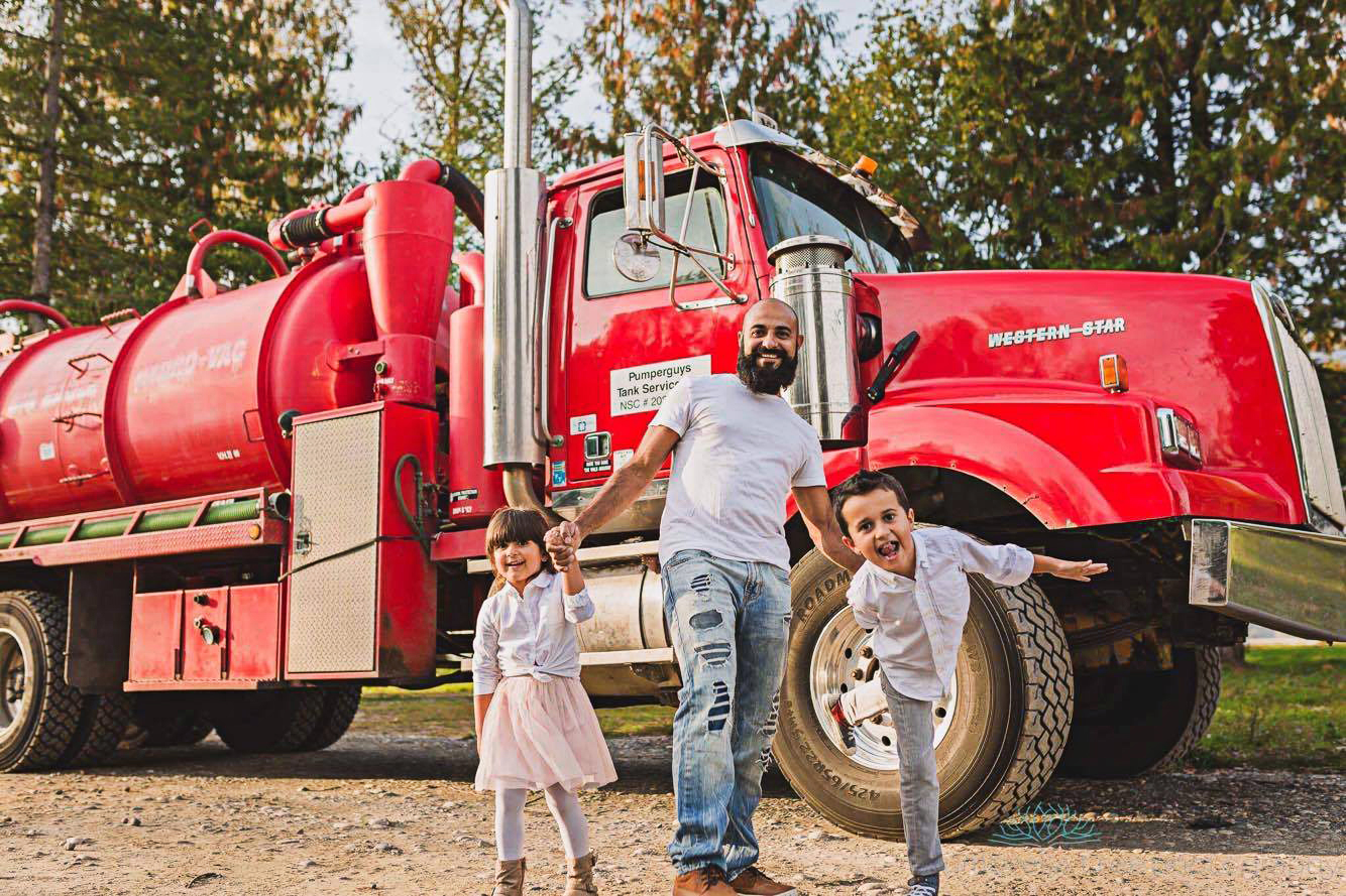 A man and two children stand smiling in front of a large, bright red Western-Star septic truck labeled "Pumperguys Tank Service" in Coquitlam BC.