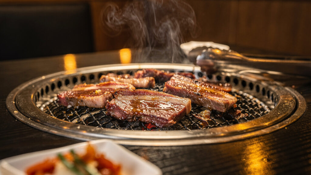 Korean BBQ charcoal grill with marbled beef slices sizzling on a table, smoke rising, tongs beside the grill