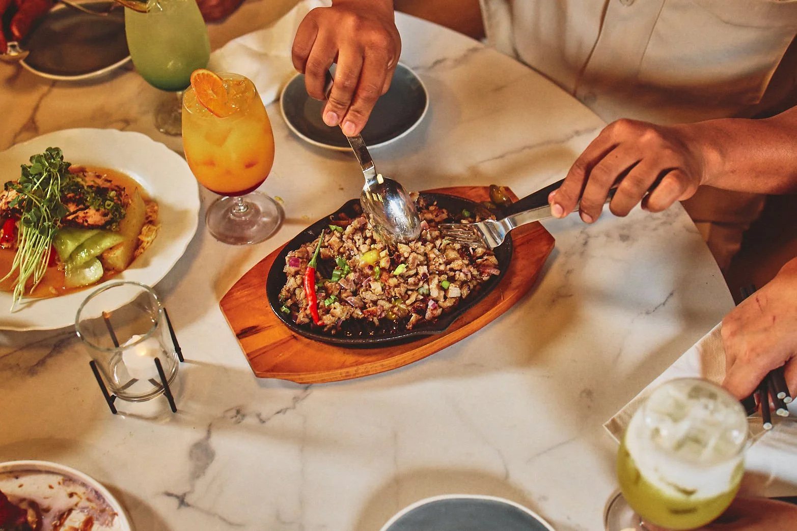 Diners reaching for Sizzling Sisig on a hot plate at Halina, with cocktails on the table